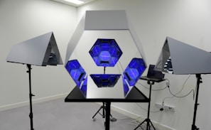 A hexagonal mirror structure is flanked by two triangular mirrors on stands, with blue geometric patterns inside. The setup is in a room with a laptop connected to the mirrors.