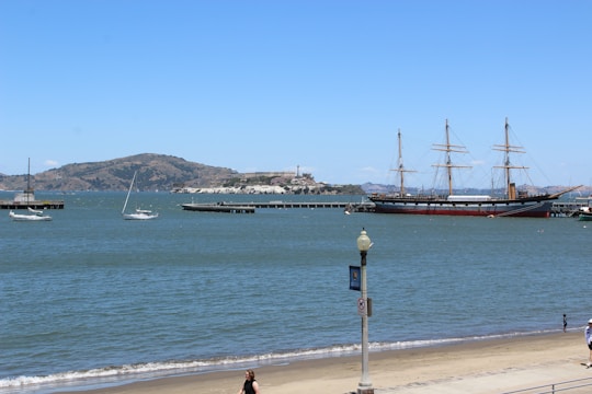 A scenic coastal view featuring calm blue waters with a few sailboats and a large historic three-masted ship docked near a pier. In the background, there are hills and a distant structure on an island. The foreground shows a sandy beach with a few people walking, and a lamppost near the edge of the water.