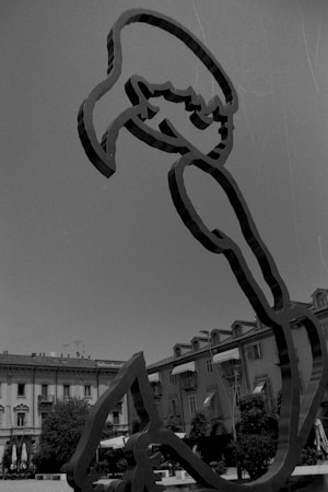 A black and white image featuring an abstract metal sculpture in the foreground, positioned in a plaza. The sculpture appears to be outline-based, possibly depicting a human form in an artistic manner. In the background, there are several buildings with colonial architecture, including balconies and windows. There are also trees and umbrellas from outdoor seating areas visible below the sculpture.