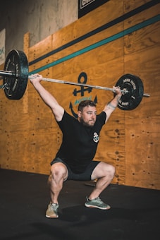 A person is performing a weightlifting exercise, holding a barbell overhead while squatting. The background features wooden paneling with an anchor logo and gym-related signage. The individual is wearing a black shirt and shorts with sneakers.