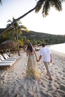 A couple is walking hand in hand along a sandy beach lined with lounge chairs and palm trees. The woman is wearing a floral dress and the man is in a white shirt and khaki pants. The sun appears to be setting, casting a warm glow over the scene, while hills with buildings overlook the beach.
