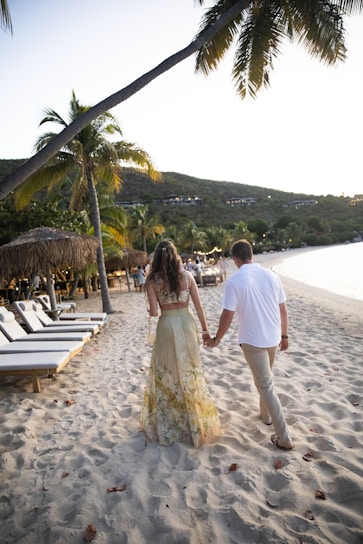 A couple is walking hand in hand along a sandy beach lined with lounge chairs and palm trees. The woman is wearing a floral dress and the man is in a white shirt and khaki pants. The sun appears to be setting, casting a warm glow over the scene, while hills with buildings overlook the beach.