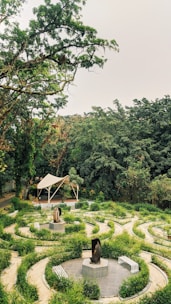 A lush green garden with a circular pathway design, featuring art sculptures in the center of the spirals. The garden is surrounded by tall, dense trees, giving it a serene and secluded atmosphere. A modern canopy structure is visible in the background, adding a touch of architecture to the natural setting.