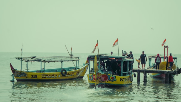 Several colorful boats are docked by a wooden pier. People, some with children, can be seen on the pier and inside the boats. Each boat carries a few small flags. The scene is set against a calm body of water under an overcast sky.