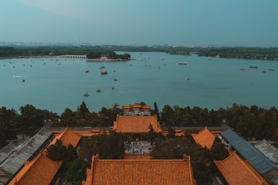 A tranquil scene featuring a large lake with many small boats scattered across its surface. In the foreground, traditional Chinese architecture with distinct orange-tiled roofs is surrounded by lush greenery. The background presents a panoramic view of a cityscape under a hazy sky, with a long bridge seen in the distance.