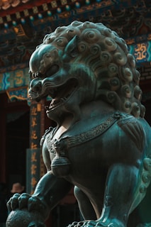 A detailed bronze statue of a mythical lion-like creature with elaborate curls in its mane and ornate decorations adorning its body. The background features traditional architecture with intricate patterns and Chinese characters.