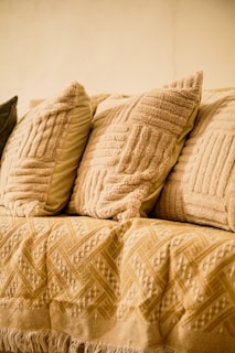 A cozy arrangement of textured throw pillows in varying shades of beige and cream is placed on a sofa. The fabric of the pillows has a detailed pattern, adding to the warm and inviting atmosphere. A woven blanket with intricate geometric designs is draped over the sofa, enhancing the comfortable setting.