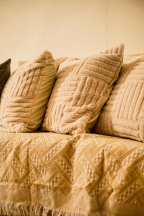 A cozy arrangement of textured throw pillows in varying shades of beige and cream is placed on a sofa. The fabric of the pillows has a detailed pattern, adding to the warm and inviting atmosphere. A woven blanket with intricate geometric designs is draped over the sofa, enhancing the comfortable setting.