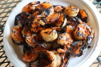 A plate filled with grilled shrimp, characterized by a charred exterior and a brightly cooked orange color. The shrimp are piled on a white plate with noticeable grill marks, suggesting they have been cooked on an open flame.