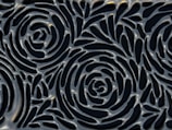 A textured surface featuring an intricate pattern of interconnected spirals reminiscent of rose flowers. The design is metallic with a glossy finish, highlighting curves and depth in the pattern.