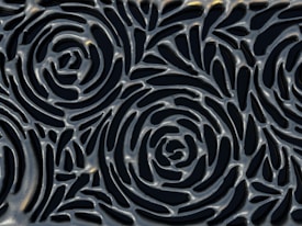 A textured surface featuring an intricate pattern of interconnected spirals reminiscent of rose flowers. The design is metallic with a glossy finish, highlighting curves and depth in the pattern.