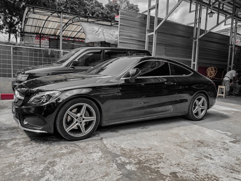 A sleek black coupe car is parked in an indoor garage setting. The structure has a metal framework with some signage visible in the background. Another vehicle is parked nearby, and there are some items such as a rolled-up hose and a small stool on the right side.
