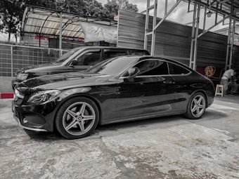 A sleek black coupe car is parked in an indoor garage setting. The structure has a metal framework with some signage visible in the background. Another vehicle is parked nearby, and there are some items such as a rolled-up hose and a small stool on the right side.