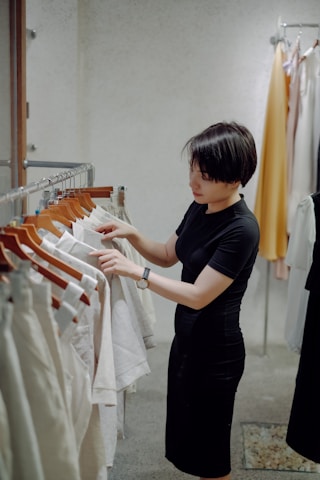 A person with short hair wearing a black dress is browsing through a clothing rack filled with light-colored garments. The setting appears to be a modern clothing store with a minimalist design.
