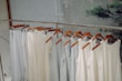 A row of light-colored sleeveless tops and pants hang neatly on wooden hangers. The clothing is displayed on a metal rack in a minimalist setting with a calm, natural background.