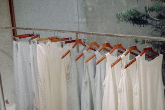 A row of light-colored sleeveless tops and pants hang neatly on wooden hangers. The clothing is displayed on a metal rack in a minimalist setting with a calm, natural background.