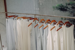 A row of light-colored sleeveless tops and pants hang neatly on wooden hangers. The clothing is displayed on a metal rack in a minimalist setting with a calm, natural background.
