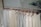 A row of light-colored sleeveless tops and pants hang neatly on wooden hangers. The clothing is displayed on a metal rack in a minimalist setting with a calm, natural background.