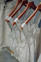 Neutral-colored garments, including dresses and pants, hang on wooden hangers. The clothing appears to be made of linen or a similar fabric, suggesting a casual and natural look.