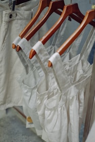 Neutral-colored garments, including dresses and pants, hang on wooden hangers. The clothing appears to be made of linen or a similar fabric, suggesting a casual and natural look.