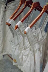 Neutral-colored garments, including dresses and pants, hang on wooden hangers. The clothing appears to be made of linen or a similar fabric, suggesting a casual and natural look.