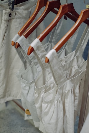 Neutral-colored garments, including dresses and pants, hang on wooden hangers. The clothing appears to be made of linen or a similar fabric, suggesting a casual and natural look.