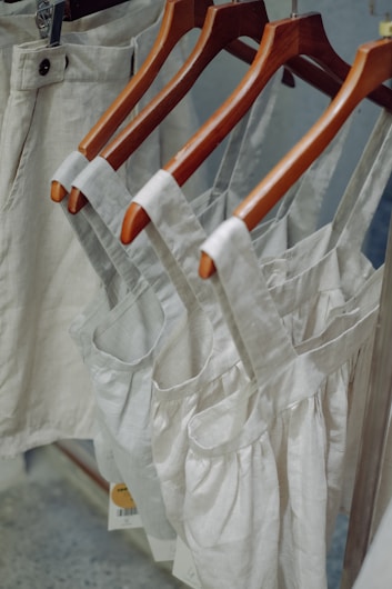 Neutral-colored garments, including dresses and pants, hang on wooden hangers. The clothing appears to be made of linen or a similar fabric, suggesting a casual and natural look.
