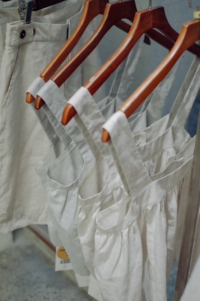 Neutral-colored garments, including dresses and pants, hang on wooden hangers. The clothing appears to be made of linen or a similar fabric, suggesting a casual and natural look.