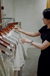 a woman in a black dress looking at a rack of white shirts