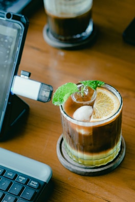 A glass filled with a layered iced coffee sits on a wooden table. The drink is garnished with a slice of orange, a mint sprig, and a scoop of ice cream. Nearby, part of a keyboard and a tablet stand are visible, suggesting a workspace setup.