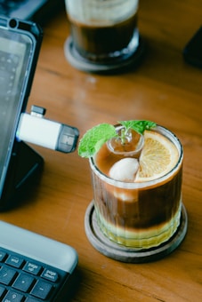 A glass filled with a layered iced coffee sits on a wooden table. The drink is garnished with a slice of orange, a mint sprig, and a scoop of ice cream. Nearby, part of a keyboard and a tablet stand are visible, suggesting a workspace setup.