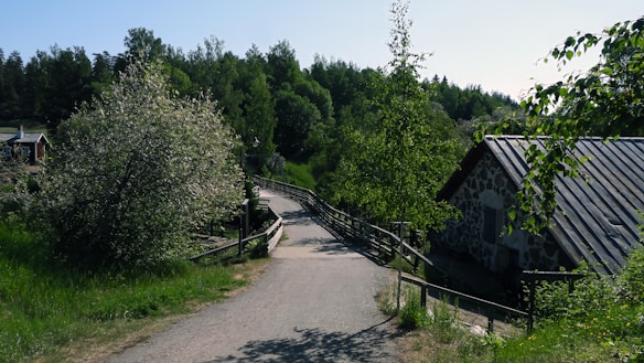 A picturesque rural scene features a narrow, winding path bordered by a wooden fence. Lush greenery and trees surround the path, with a stone cottage visible on the right. A mix of shaded and sunlit areas adds depth to the landscape, creating a serene and peaceful environment.