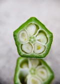 A close-up cross-section of a fresh green okra pod, revealing the intricate interior structure with small white seeds and a soft, fleshy core surrounded by a crisp outer layer. The focus on the texture and details showcases the natural patterns found in the vegetable.