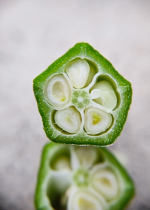 A close-up cross-section of a fresh green okra pod, revealing the intricate interior structure with small white seeds and a soft, fleshy core surrounded by a crisp outer layer. The focus on the texture and details showcases the natural patterns found in the vegetable.