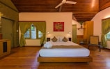 A well-furnished bedroom with a wooden ceiling and floor. The room features a bed with white and beige bedding, flanked by two nightstands with small lamps. There are green curtains on the windows, and a vibrant red painting hangs above the bed. A potted plant sits on a wooden desk in the corner, and a wicker chair is present nearby, adding a natural touch to the decor.