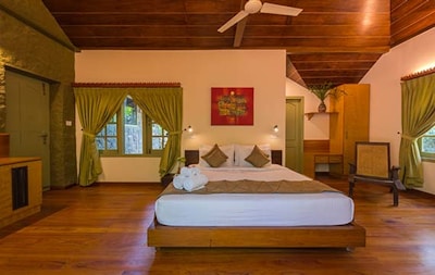 A well-furnished bedroom with a wooden ceiling and floor. The room features a bed with white and beige bedding, flanked by two nightstands with small lamps. There are green curtains on the windows, and a vibrant red painting hangs above the bed. A potted plant sits on a wooden desk in the corner, and a wicker chair is present nearby, adding a natural touch to the decor.