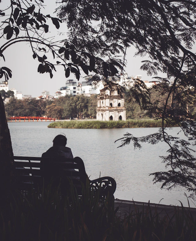 A person sits on a lakeside bench looking across Hoan Kiem Lake toward the ancient Turtle Tower rising from the water.