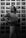 A person stands confidently in front of a wall displaying numerous black and white photographs, holding a camera. The photographs depict various scenes and people, suggesting a gallery or personal exhibition.