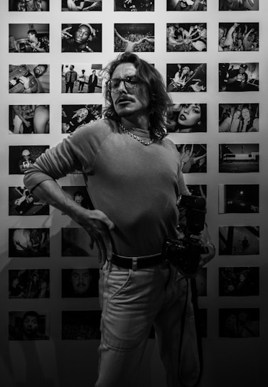 A person stands confidently in front of a wall displaying numerous black and white photographs, holding a camera. The photographs depict various scenes and people, suggesting a gallery or personal exhibition.