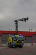 A large, green firefighting vehicle with multiple axles is stationed in front of a building with red trim and large garage doors. A tall lift arm extends from the top of the vehicle, equipped with various nozzles and equipment. The sky is overcast, suggesting a cloudy day.