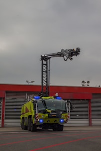 A large, green firefighting vehicle with multiple axles is stationed in front of a building with red trim and large garage doors. A tall lift arm extends from the top of the vehicle, equipped with various nozzles and equipment. The sky is overcast, suggesting a cloudy day.