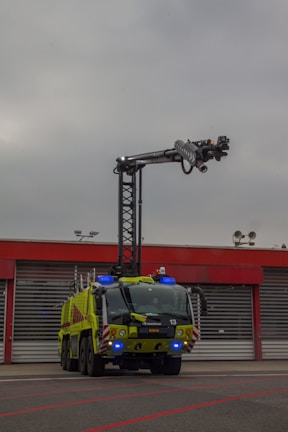A large, green firefighting vehicle with multiple axles is stationed in front of a building with red trim and large garage doors. A tall lift arm extends from the top of the vehicle, equipped with various nozzles and equipment. The sky is overcast, suggesting a cloudy day.