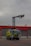 A large, green firefighting vehicle with multiple axles is stationed in front of a building with red trim and large garage doors. A tall lift arm extends from the top of the vehicle, equipped with various nozzles and equipment. The sky is overcast, suggesting a cloudy day.