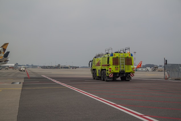An airport scene featuring a bright green airport fire truck driving on the tarmac. Various aircraft tails are visible in the background, with the sky overcast. The foreground is marked with multiple red and white lines on the pavement.