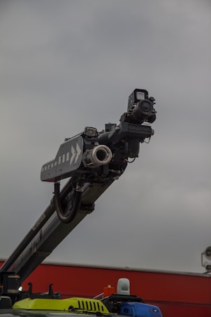 A large, industrial piece of machinery with a mechanical arm and attachments pointing upward, set against a cloudy sky. The machinery is mounted on a colorful base that includes yellow and blue colors.