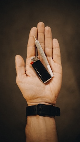 A close-up of a hand holding a lighter and a rolled cigarette. The hand is positioned palm-up, with the lighter and cigarette resting on it. The background is a neutral, blurred surface, and the person is wearing a black wristband.