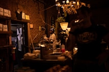 The image depicts a dimly lit interior of a coffee shop or caf&eacute; with a brick wall. A man is standing near coffee-making equipment, wearing gloves and smiling. A chandelier with candles hangs from the ceiling. Another person is visible in the foreground, partially silhouetted. Coffee bags and various equipment are visible, suggesting a cozy and warm atmosphere.