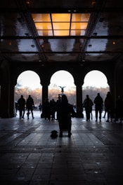 A silhouette of a musician playing an instrument under a beautifully designed ceiling with light reflections. People are walking in the background through an archway, leading to an open park area with a central fountain.