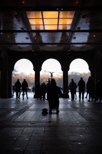 A silhouette of a musician playing an instrument under a beautifully designed ceiling with light reflections. People are walking in the background through an archway, leading to an open park area with a central fountain.