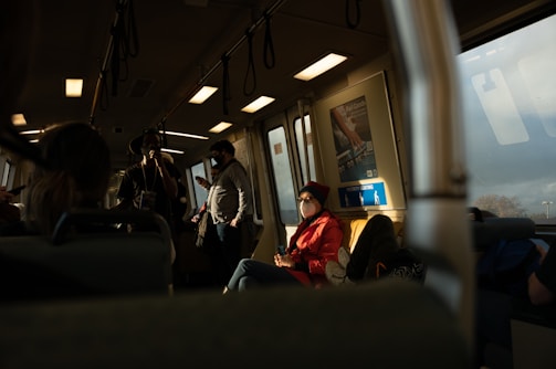 Passengers inside a dimly lit train carriage, with some standing and others sitting. Sunlight streams through a window, casting shadows and illuminating a person wearing a mask and red jacket.